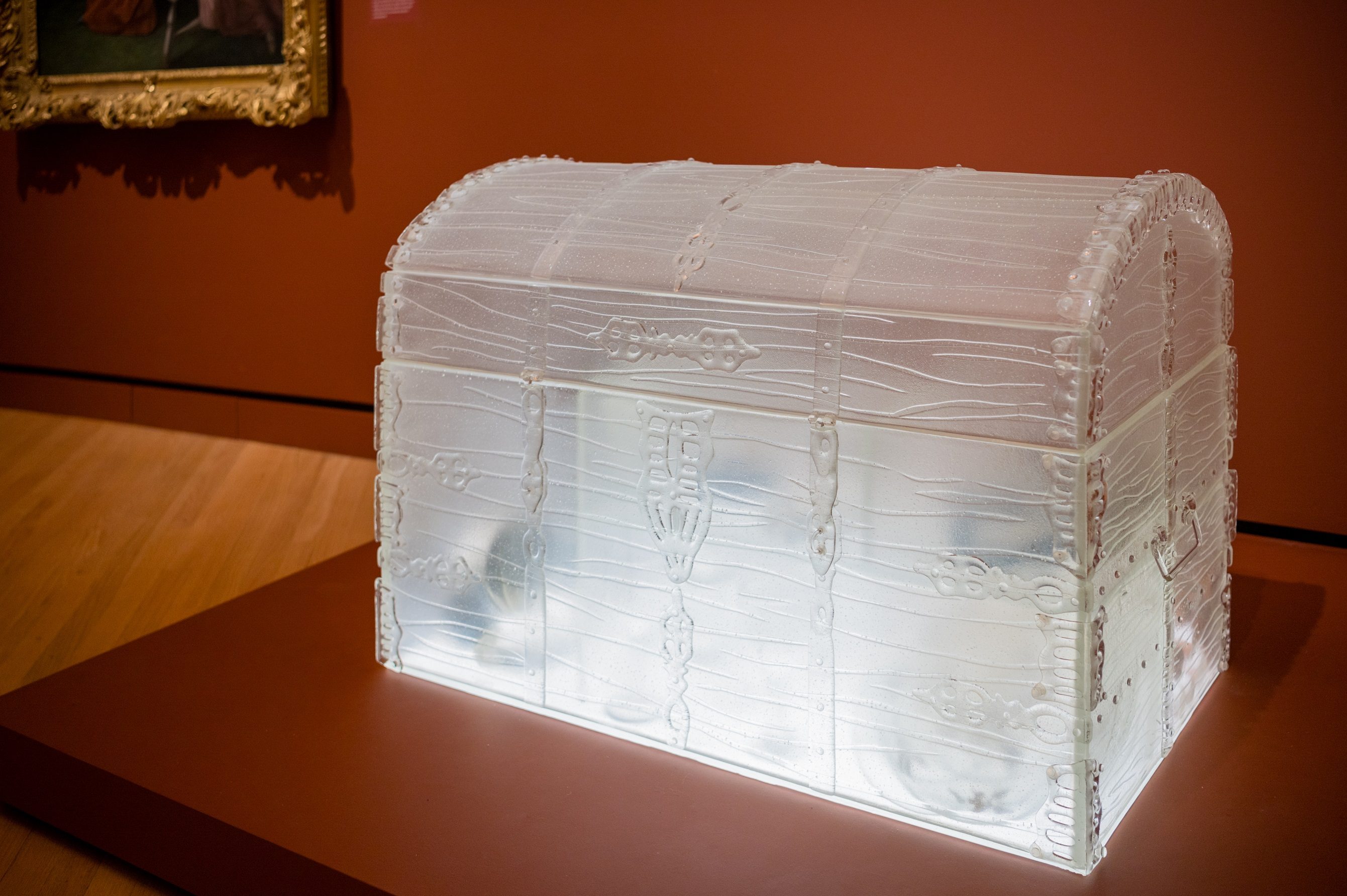Beth Lipman, Belonging(s), 2020, glass, ceramic, gold lacquer, enamel paint, salt, sand, and adhesive, 27 in. × 40 1/2 in. × 23 in. Crystal Bridges Museum of American Art, Bentonville, Arkansas, 2021.3. Photo by Ironside Photography. Transparent glass treasure chest with intricate designs in a museum, painting in background.