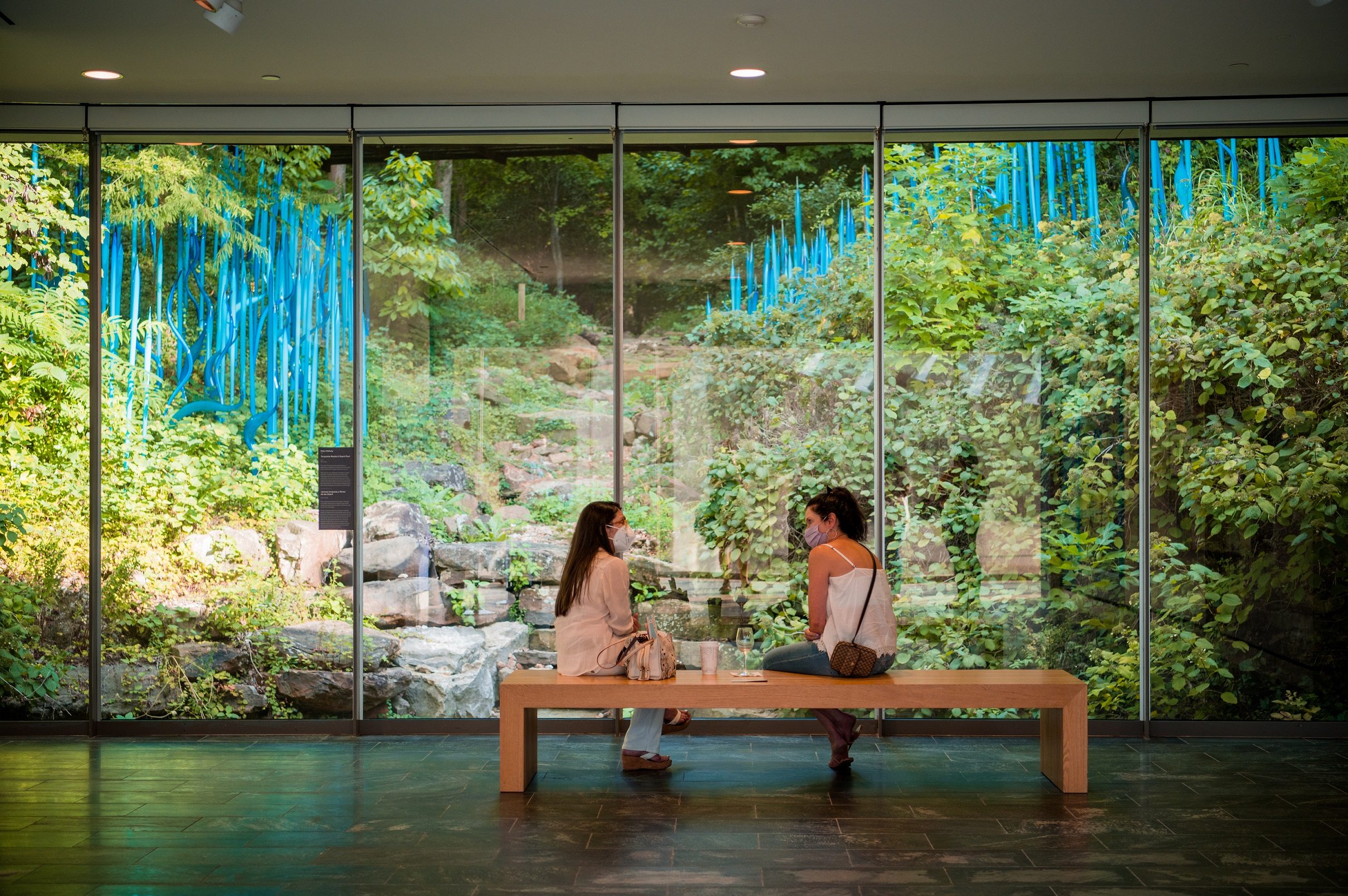 Guests sitting in front of of a glass window looking out to a Chihuly artwork Photo by Ironside Photography / Stephen Ironside. two guests sit on a bench in front of a glass window looking out to a Chihuly artwork