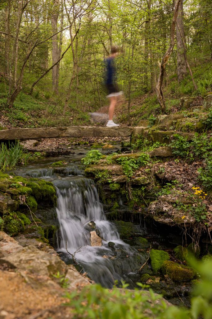 Blurred person crossing narrow stone bridge over small waterfall in lush green forest.