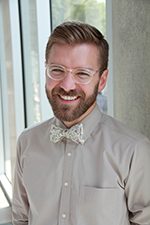 symposium-hs-alligood-chad Man with glasses, beard, bow tie, smiling indoors near large windows in a long-sleeved shirt.