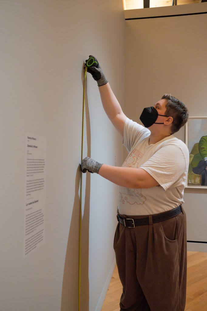 Person in mask and gloves measures gallery wall near artworks and information plaques.