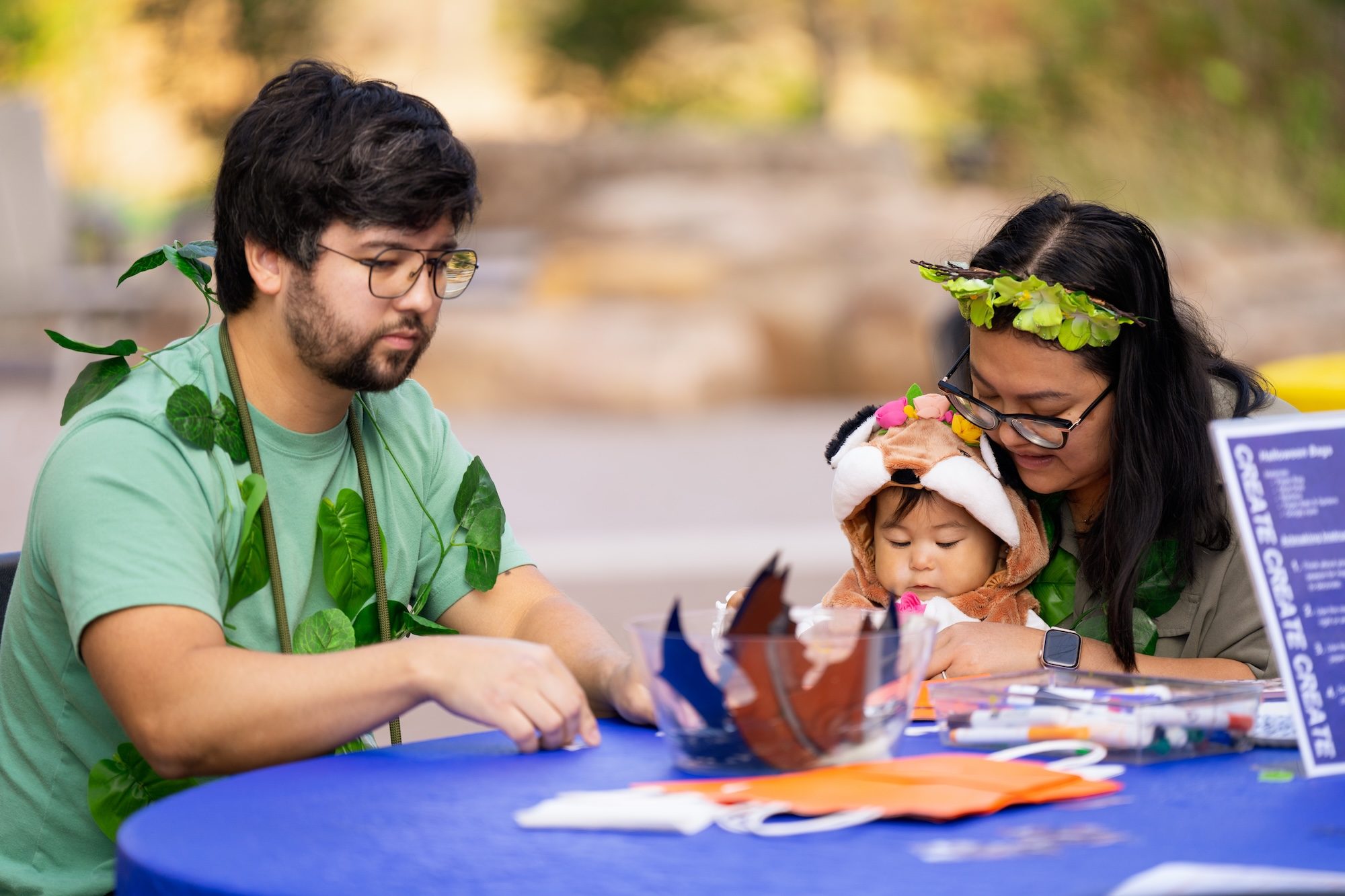 Fall family artmaking Adults with foliage accessories and child in costume crafting at blue table with art supplies.