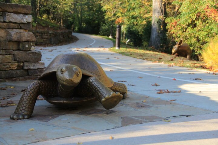 Nancy Schön, Tortoise and Hare, 2009, silicon bronze, Tortoise: 29 x 29 x 60 in., Hare: 36 x 24 x 29 in. Crystal Bridges Museum of American Art, Bentonville, Arkansas, 2009.8 Bronze turtle statue on a paved path with a bronze hare in the background in a lush, green landscape.
