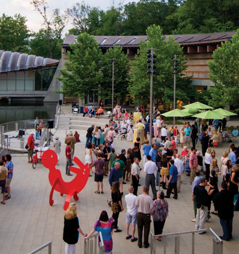 Party Outdoor event on patio with diverse crowd, red sculpture, tables, umbrellas, surrounded by trees.